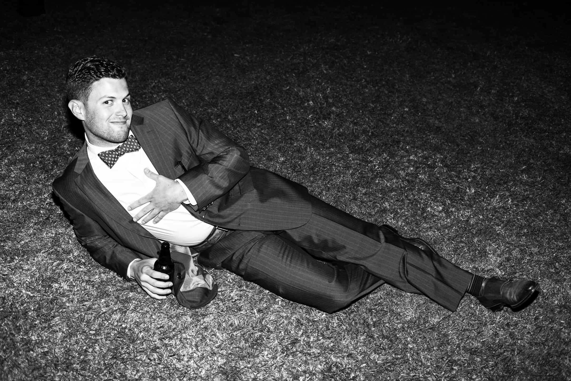 man with bow tie lying down in the grass doing funny pose and drinking a beer